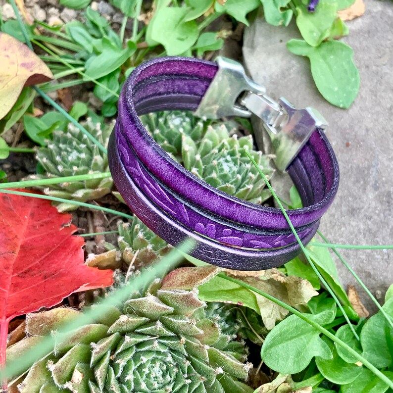 Pu&ograve; includere: Un braccialetto in pelle viola con una chiusura in argento. Il braccialetto &egrave; composto da due fili di pelle e presenta un motivo floreale.