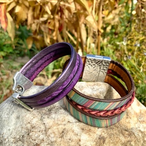 Pu&ograve; includere: Due braccialetti in pelle, uno viola con chiusura argentata e l'altro con un design intrecciato in tonalit&agrave; di marrone, verde e rosa.