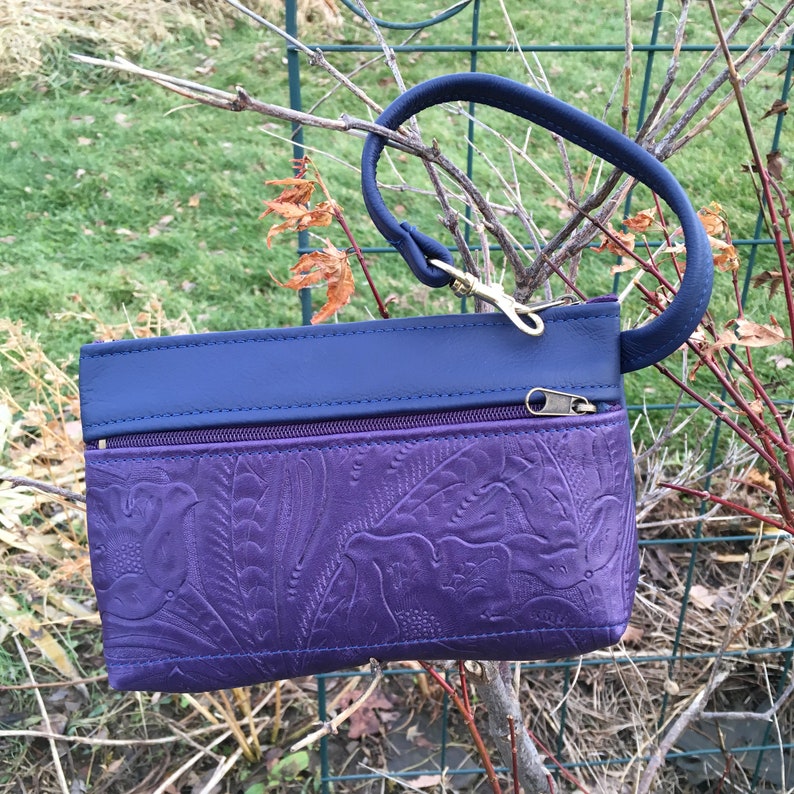 May include: A small purple leather clutch with a navy blue top flap and a navy blue wrist strap. The clutch has a zipper closure and an embossed floral design.
