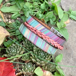 Pu&ograve; includere: Un braccialetto in pelle multicolore con un cinturino in pelle intrecciata rosa. Il braccialetto &egrave; realizzato in pelle a righe con sfumature di rosa, blu e verde.