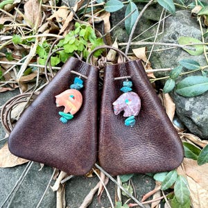 Puede incluir: Dos bolsas de cuero marrón oscuro con cierre de cordón. Cada bolsa tiene un colgante de oso tallado con turquesa y otros acentos de piedra. Las bolsas se exhiben sobre un lecho de hojas y vegetación.