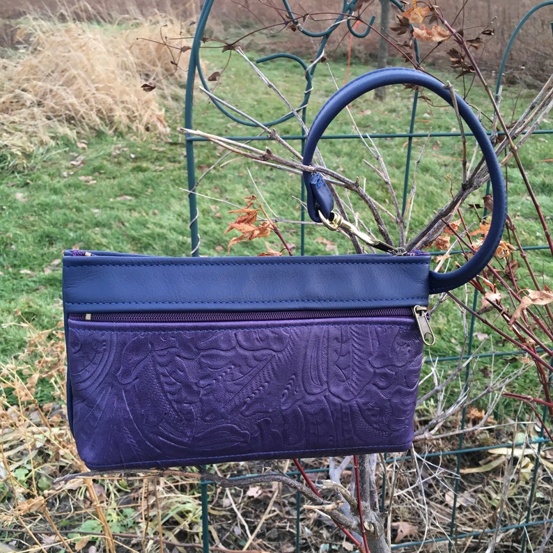 May include: A purple leather clutch with a blue leather top and a blue leather loop handle. The clutch has a zipper closure and two zippered pockets.