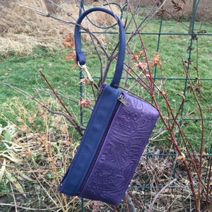 May include: A purple leather clutch with a blue leather flap and a gold zipper. The clutch has an embossed floral pattern and a blue strap.