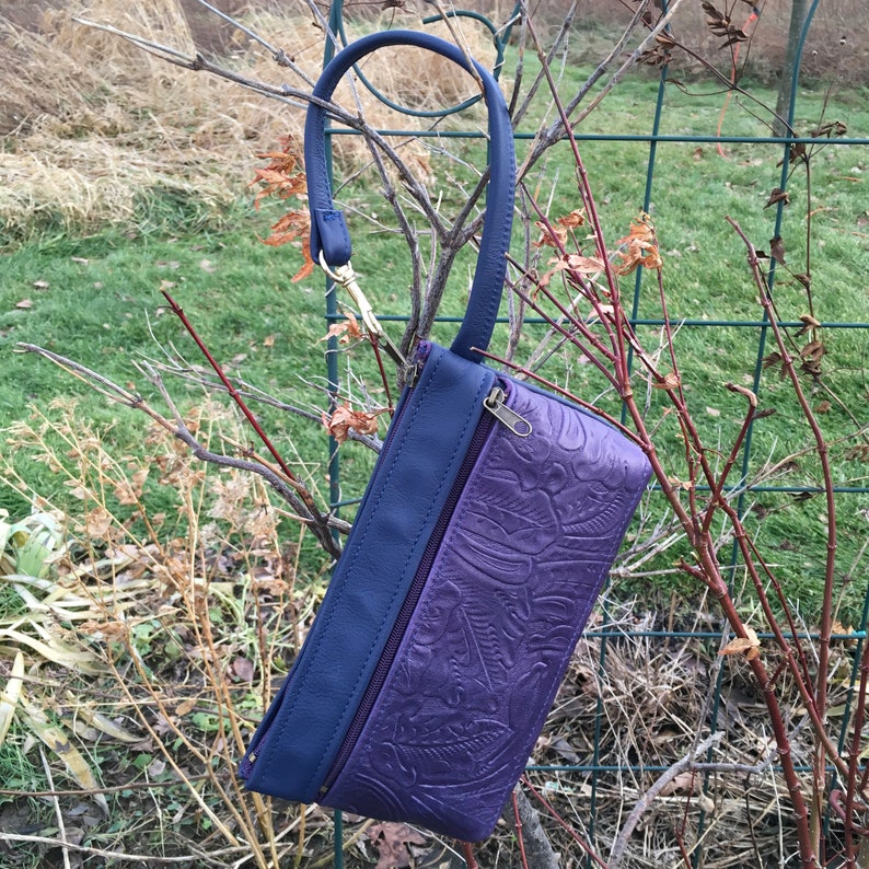 May include: A purple leather clutch with a blue leather flap and a gold zipper. The clutch has an embossed floral pattern and a blue strap.
