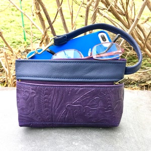 May include: A small purple leather purse with a blue leather flap and a zipper closure. The purse has a floral embossed design and is shown with a blue phone and glasses inside.
