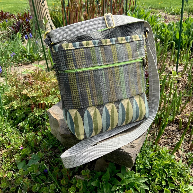 Op de afbeelding: Een grijze en groene crossbody tas met een ritsvak en een lange verstelbare schouderband.