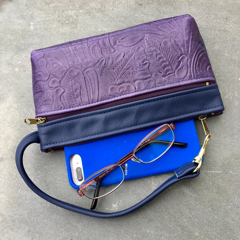 May include: A purple leather clutch with a navy blue strap and zipper closure. The clutch is open and a blue phone case with reading glasses is inside.