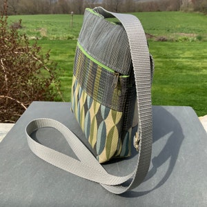 Op de afbeelding: Een grijze en groene crossbody tas met een grijze verstelbare schouderband. De tas heeft een ritssluiting en een klein vakje aan de voorkant.