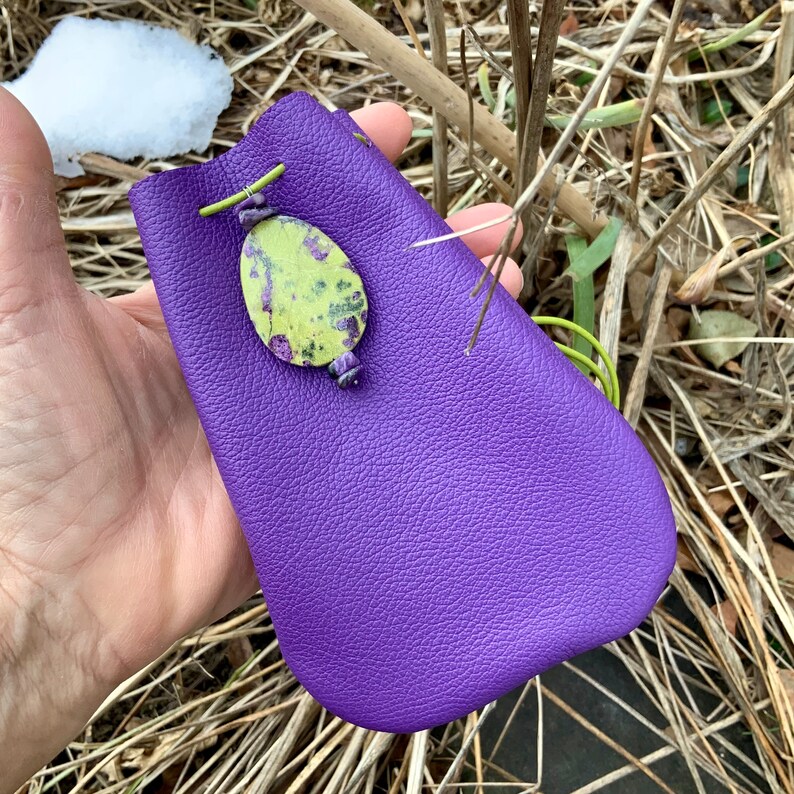 May include: A purple leather pouch with a green and yellow stone accent. The pouch is closed with a drawstring and has a green leather strap.
