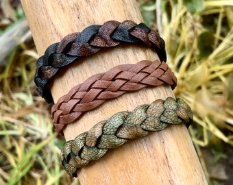 Può includere: Tre braccialetti in pelle intrecciata in diversi colori: nero e marrone, marrone e verde e marrone. I braccialetti sono esposti su una superficie in legno.