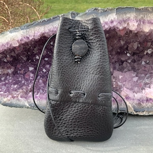 May include: A black leather pouch with a drawstring closure and a decorative wooden bead accent. The pouch is sitting on a purple and white amethyst geode.