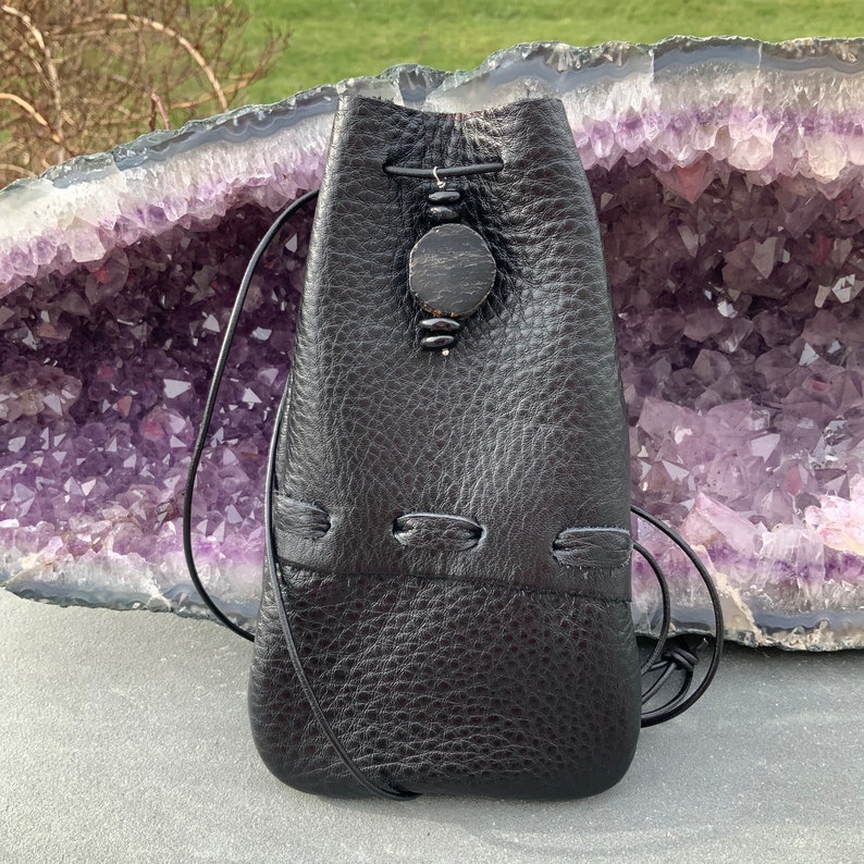 K&ouml;nnte beinhalten: Ein schwarzer Lederbeutel mit Kordelzug und einem dekorativen Holzperlen-Akzent. Der Beutel liegt auf einem violetten und wei&szlig;en Amethyst-Geoden.