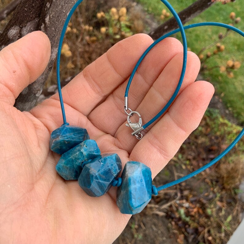 May include: A necklace featuring five faceted, blue-green gemstones strung on a teal cord. The gemstones have a rough, natural appearance. The necklace is secured with a silver-colored clasp.