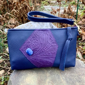 May include: A navy blue leather clutch with a purple, floral-embossed diamond-shaped accent. A blue gemstone is centered on the purple panel. The clutch has a wrist strap and a gold-colored zipper.