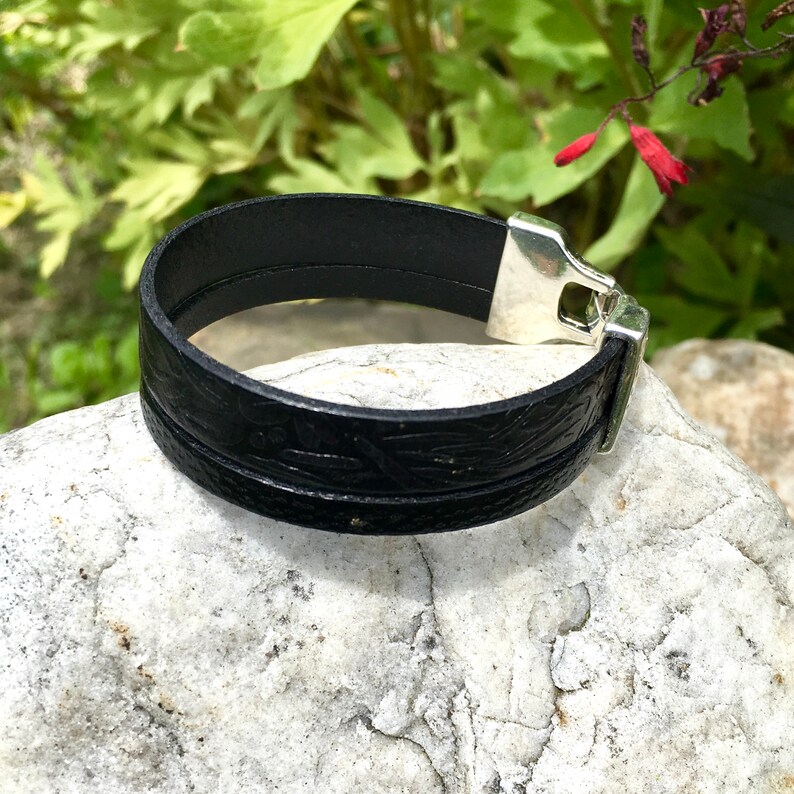 May include: A black leather bracelet with a silver clasp. The bracelet has a textured design.