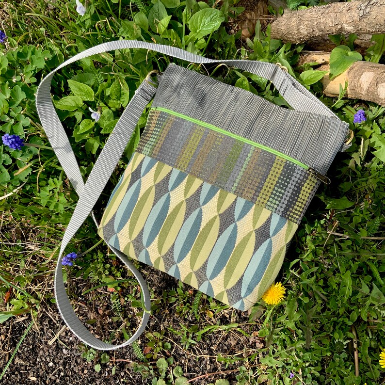 Op de afbeelding: Een grijze en groene crossbody tas met een lange verstelbare schouderband. De tas heeft een ritssluiting en een klep met een magneetsluiting.