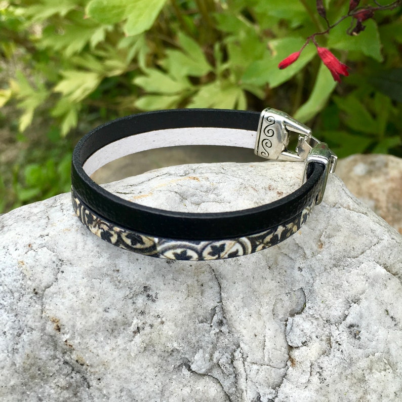 May include: A black leather bracelet with a white and black patterned design. The bracelet has a silver clasp with a decorative design.