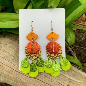 May include: A pair of dangle earrings with a layered design. The top layer is a fan-shaped piece of orange leather. The middle layer is a round piece of orange leather with a textured surface. The bottom layer is made up of five teardrop-shaped pieces of green leather.