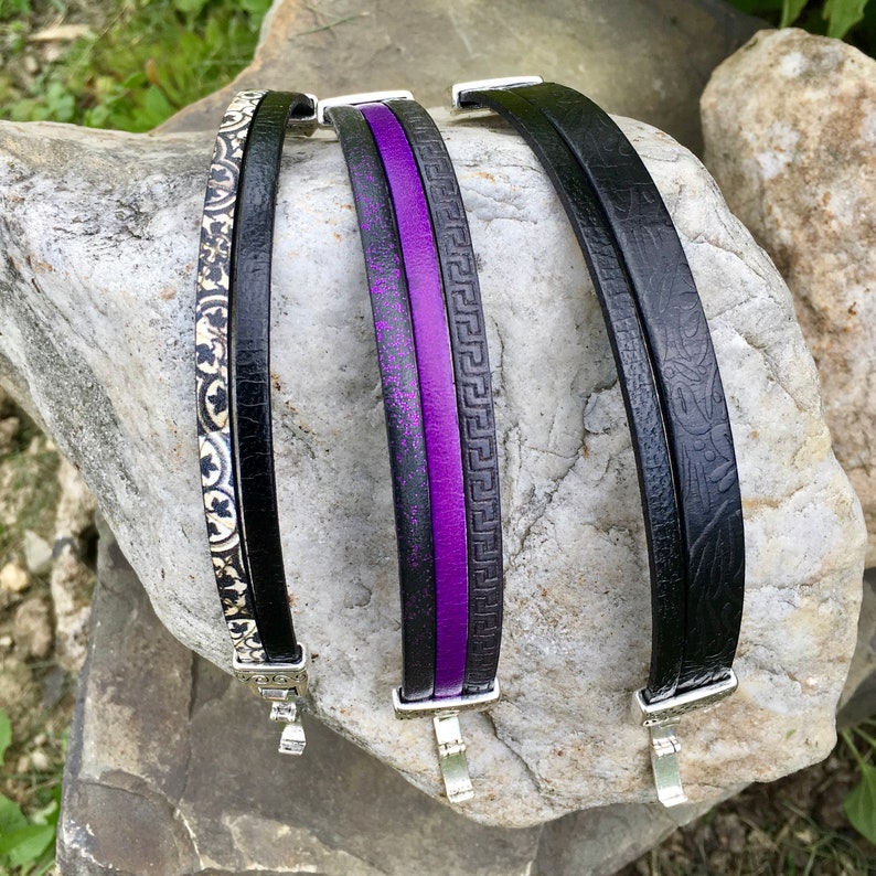 May include: Four black leather bracelets with different patterns and designs. One bracelet has a purple stripe, another has a geometric pattern, and another has a floral pattern. The bracelets are all on a grey rock surface.
