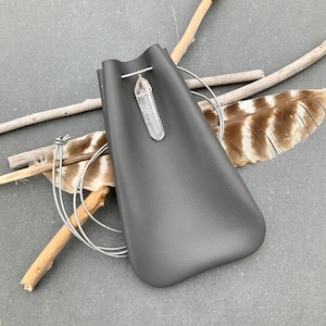 May include: A gray leather pouch with a silver chain and a clear quartz crystal pendant. The pouch is lying on a wooden surface with a feather and twigs.
