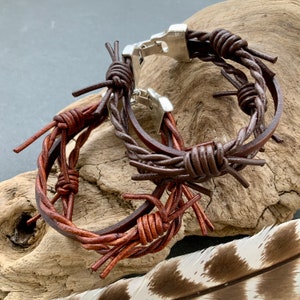 May include: A brown leather bracelet with a barbed wire design. The bracelet features a silver clasp and is made of twisted leather strands with protruding wire-like details. The bracelet is displayed on a piece of driftwood.