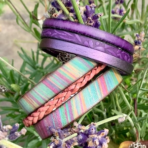 Könnte beinhalten: Ein Set aus drei Lederarmbändern. Ein Armband ist lila mit einem Blumenmuster, ein anderes ist ein geflochtenes rosa Lederarmband und das dritte ist ein mehrfarbiges gestreiftes Lederarmband.