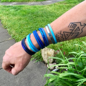 May include: A selection of blue bracelets in diverse shades, from navy to light blue and silver. Some bracelets feature textured or glittery finishes, while others are smooth. The bracelets are displayed on a wrist, presenting a variety of styles.