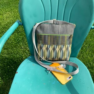 Op de afbeelding: Een groene en blauwe crossbody tas met een grijze band en een gele telefoon op een turquoise stoel.