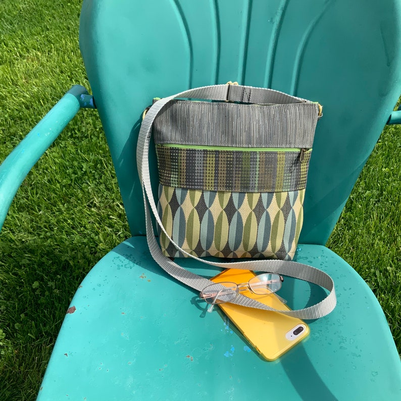 Op de afbeelding: Een groene en blauwe crossbody tas met een grijze band en een gele telefoon op een turquoise stoel.