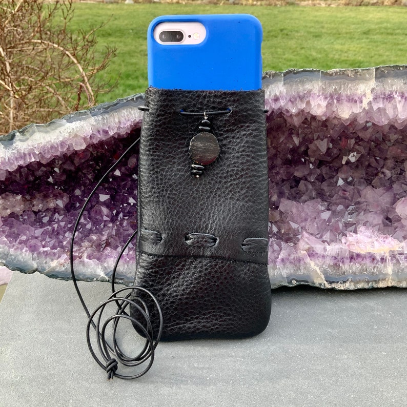 K&ouml;nnte beinhalten: Eine schwarze Leder-Handyh&uuml;lle mit einem blauen Telefon darin. Die H&uuml;lle hat eine Schnur und einen Holzperlen-Anh&auml;nger. Die H&uuml;lle liegt auf einem violetten und wei&szlig;en Kristall.