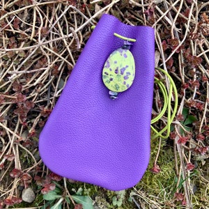 May include: A purple leather drawstring bag with a green and yellow patterned stone charm. The bag is sitting on a bed of brown twigs and green moss.