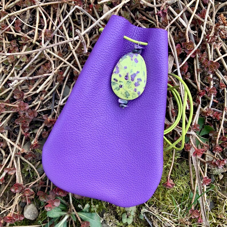 May include: A purple leather drawstring bag with a green and yellow patterned stone charm. The bag is sitting on a bed of brown twigs and green moss.