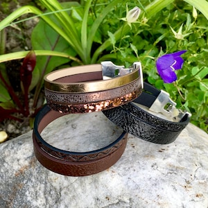 May include: Three leather bracelets stacked on a rock. The top bracelet is brown with gold and copper accents. The middle bracelet is black leather. The bottom bracelet is brown with a wavy design.