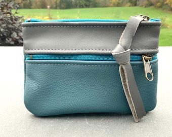 Può includere: Piccola pochette in pelle verde acqua e grigia con due scomparti con cerniera. La pochette ha una parte superiore in pelle grigia e una cerniera color verde acqua. Un cinturino in pelle grigia è attaccato alla cerniera.