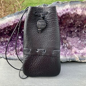 K&ouml;nnte beinhalten: Ein schwarzer Lederbeutel mit Kordelzug und einem dekorativen Holzperlen-Akzent. Der Beutel h&auml;ngt an einer violetten Amethyst-Geode.
