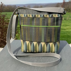 Op de afbeelding: Een grijze en groene crossbody tas met een grijze verstelbare schouderband. De tas heeft een ritsvak en een klep sluiting.
