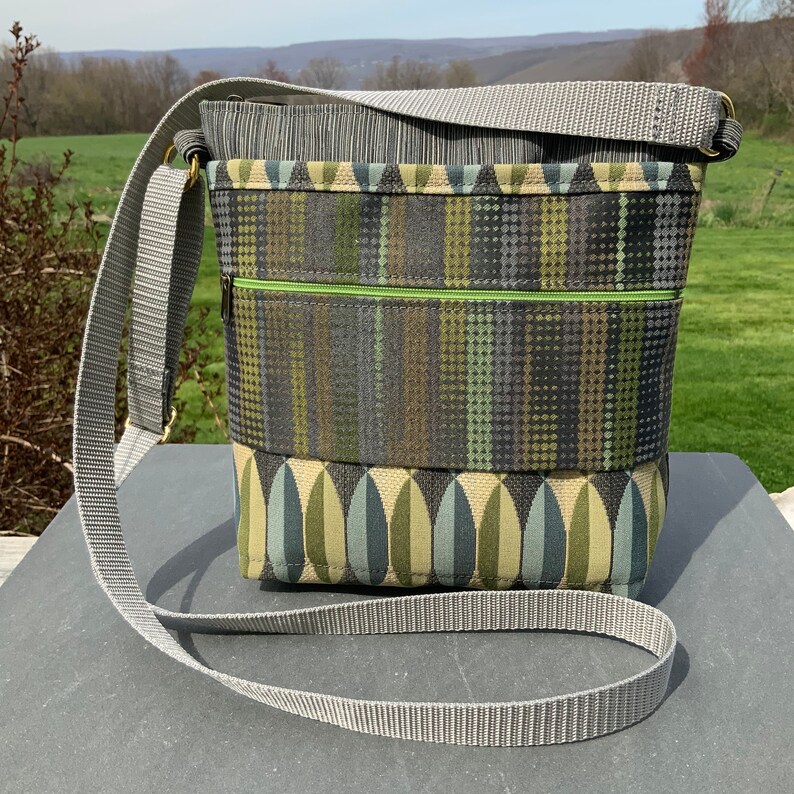 Op de afbeelding: Een grijze en groene crossbody tas met een grijze verstelbare schouderband. De tas heeft een ritsvak en een klep sluiting.