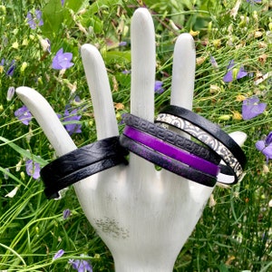 May include: A set of four bracelets, one black leather, one black with a white and silver geometric pattern, one black with a purple stripe, and one black with a white and silver floral pattern.