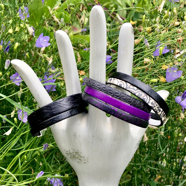 May include: A set of four bracelets, one black leather, one black with a white and silver geometric pattern, one black with a purple stripe, and one black with a white and silver floral pattern.