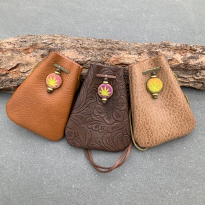 Puede incluir: Tres bolsas de cuero con cordones y dijes decorativos en forma de hoja. Las bolsas son de color marrón, beige y marrón oscuro con diseños florales en relieve.