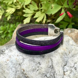 May include: A multi-layered leather bracelet with a silver clasp. The bracelet features a purple leather strap with a black leather strap with a Greek key pattern.
