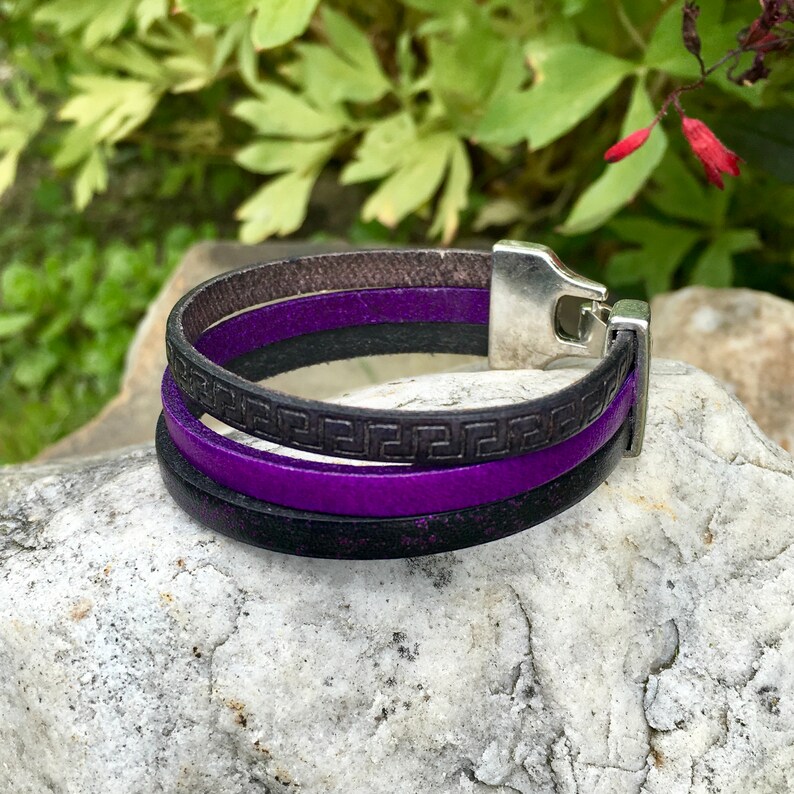 May include: A multi-layered leather bracelet with a silver clasp. The bracelet features a purple leather strap with a black leather strap with a Greek key pattern.