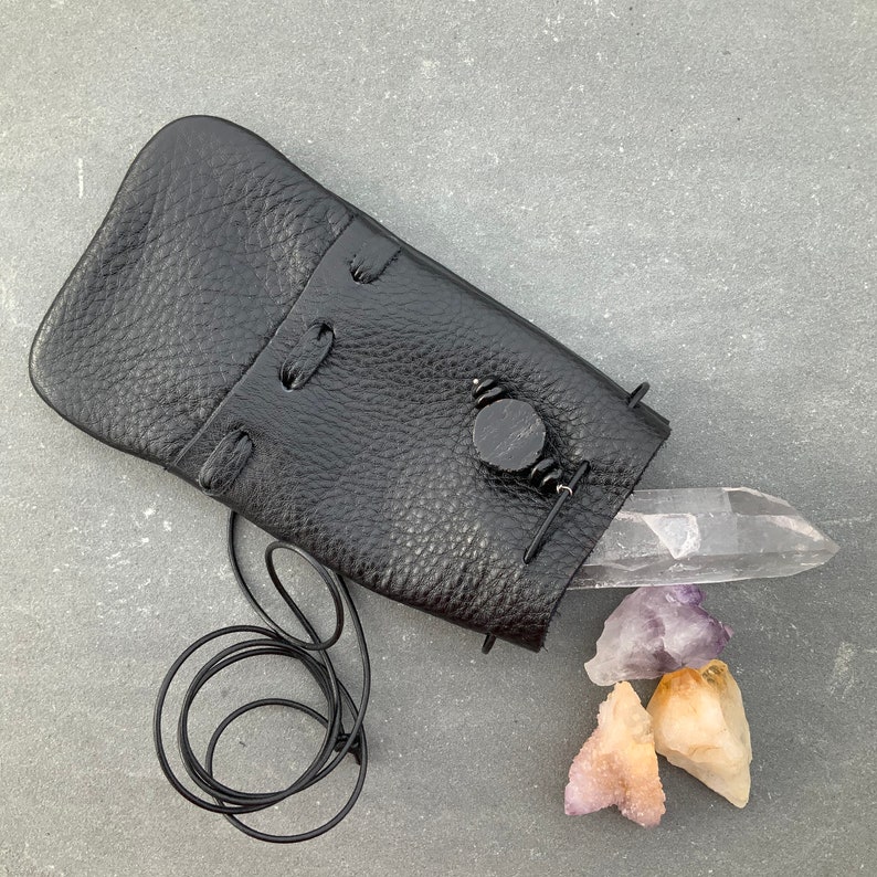 K&ouml;nnte beinhalten: Eine schwarze Ledertasche mit Kordelzug und einem kleinen schwarzen Knopf. Die Tasche ist offen und enth&auml;lt einen klaren Quarzkristall und drei Amethystkristalle.