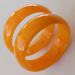 May include: Two orange bangles with a smooth, glossy finish. The bracelets are a warm, translucent orange colour, and they are stacked together. Light reflects off the curved surfaces, creating highlights and shadows.