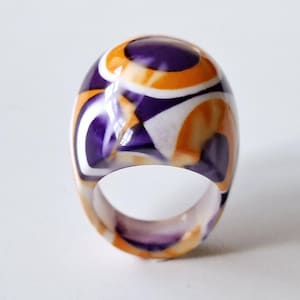 May include: A statement ring with a striking, abstract pattern. The ring showcases a swirl design in purple, orange, and white. It's made from a glossy material and has a rounded, dome-like form.