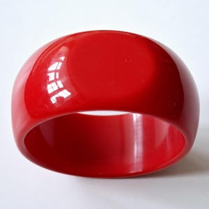 May include: A solid, bright red bangle bracelet with a smooth, glossy, and reflective surface. The circular accessory features a wide, thick band design with a uniform, bold colour throughout, showcasing a high-shine finish that catches light.