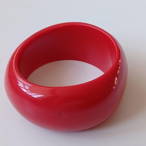 May include: A bright red bangle bracelet. The smooth, glossy surface reflects light, emphasising the rounded shape. This bold accessory is a statement piece, ideal for adding a pop of colour to any outfit. The bracelet is a solid, seamless design.