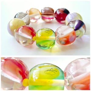 May include: A circular bracelet composed of large, spherical glass beads. Each bead features a unique swirl of colors including red, yellow, green, purple, and white. One central bead displays a thin, black, hand-drawn signature on its surface.