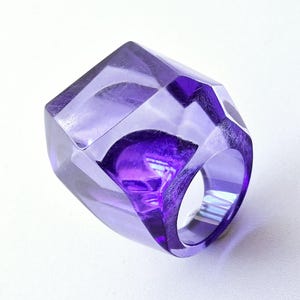 Può includere: Un anello viola traslucido caratterizzato da un design geometrico sfaccettato con molteplici superfici angolari. L'anello è realizzato in un materiale trasparente color violetto che lascia passare la luce, creando profondità e riflessi.