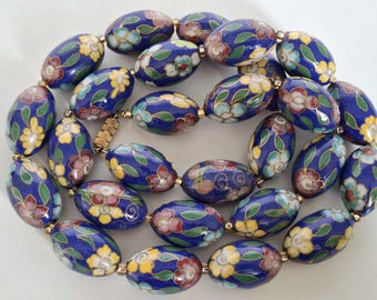 Puede incluir: Un collar con cuentas ovaladas en un tono azul oscuro, adornadas con diseños florales en amarillo, rojo y blanco. Las cuentas están unidas con conectores dorados, creando una pieza de joyería vintage.